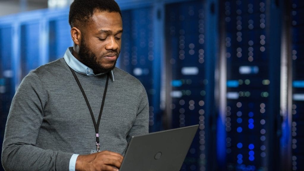 Black Data Center IT Technician Standing in Server Rack Corridor with a Laptop Computer